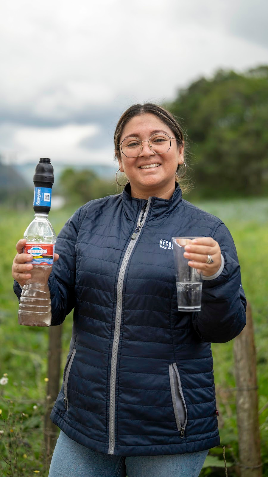 woman smiling with clean and dirty water cups, one with sawyer filter attached